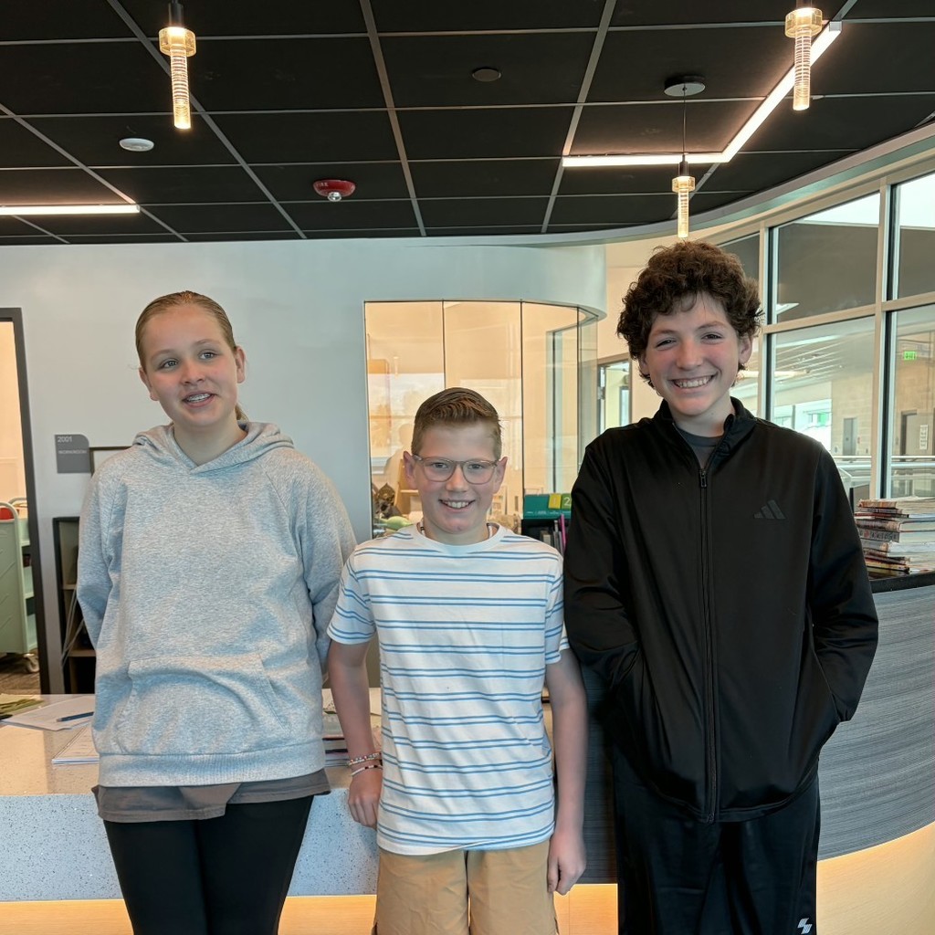 Three older elementary students smile while standing together in the school library before the District Book Battle.