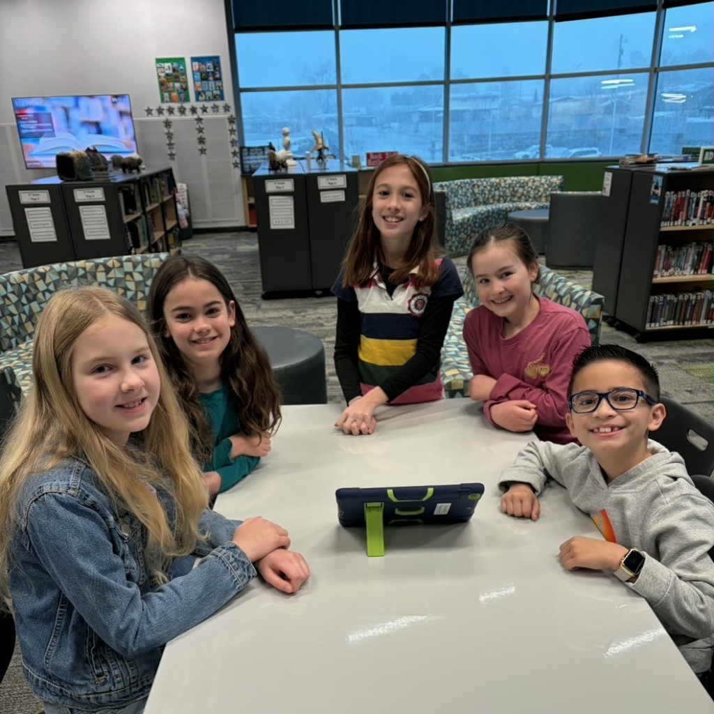 Five elementary students gather around a table with a tablet in the school library during Book Battle preparation.