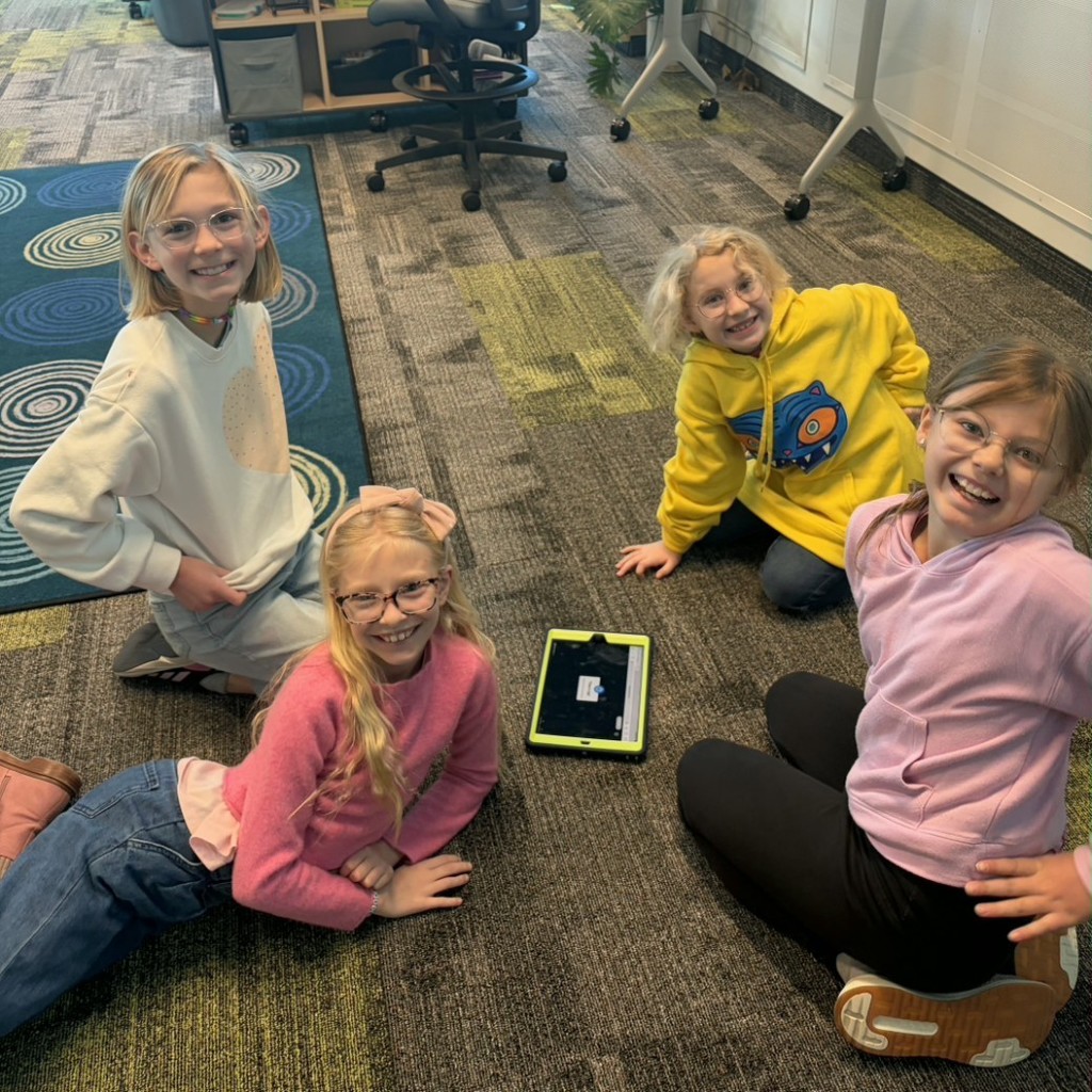 Four elementary students sit smiling on the floor around a tablet during Book Battle practice in the school library.
