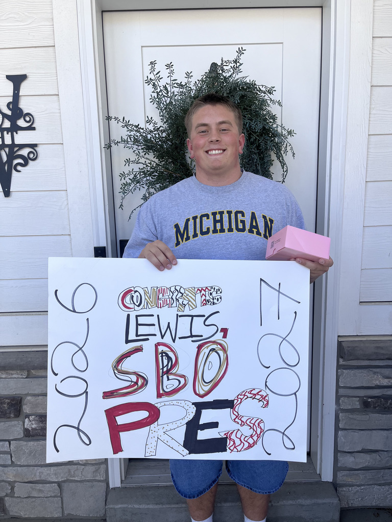 A smiling young man stands in front of a white front door holding a handmade poster that says “Congrats Lewis! SBO Pres 2026,” with red, black, and gold decorations and curly doodles along the sides. He’s wearing a gray Michigan sweatshirt and blue shorts, and holding a small pink Crumbl box.