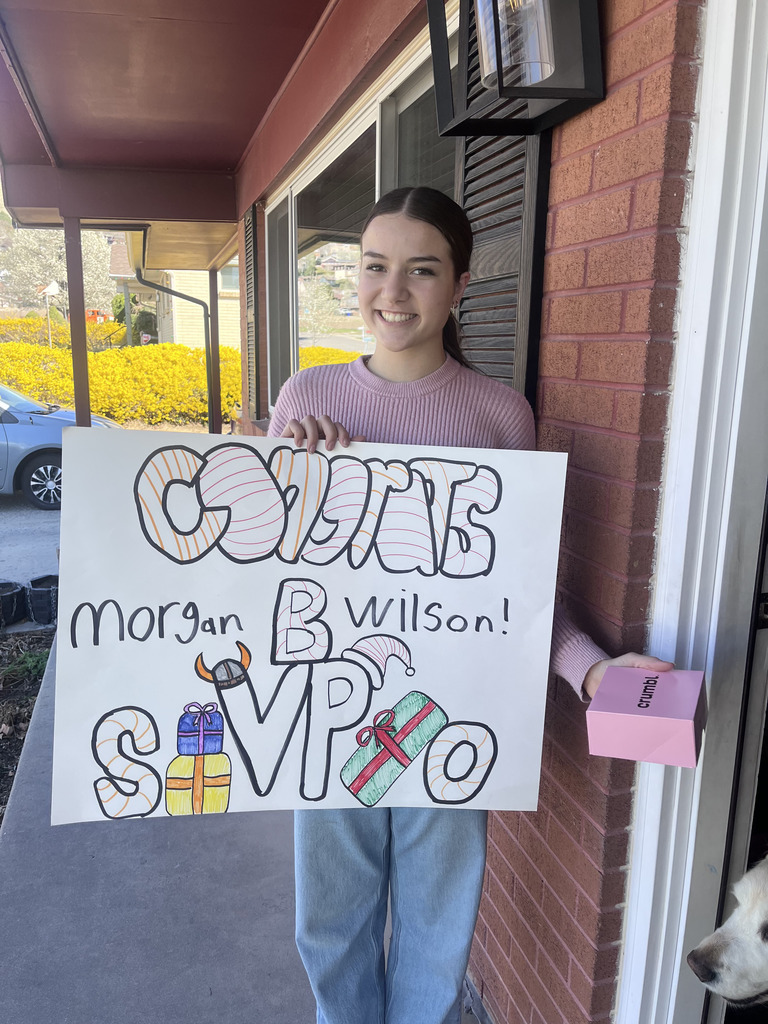 A smiling young person stands on a front porch holding a handmade poster that says “Congrats Morgan B Wilson! B SVP O,” decorated with candy-cane stripes, a Viking horn, and wrapped presents. They’re wearing a pink sweater and jeans, holding a small pink Crumbl box, with a brick wall, porch light, yellow bushes, and part of a white dog visible in the background.