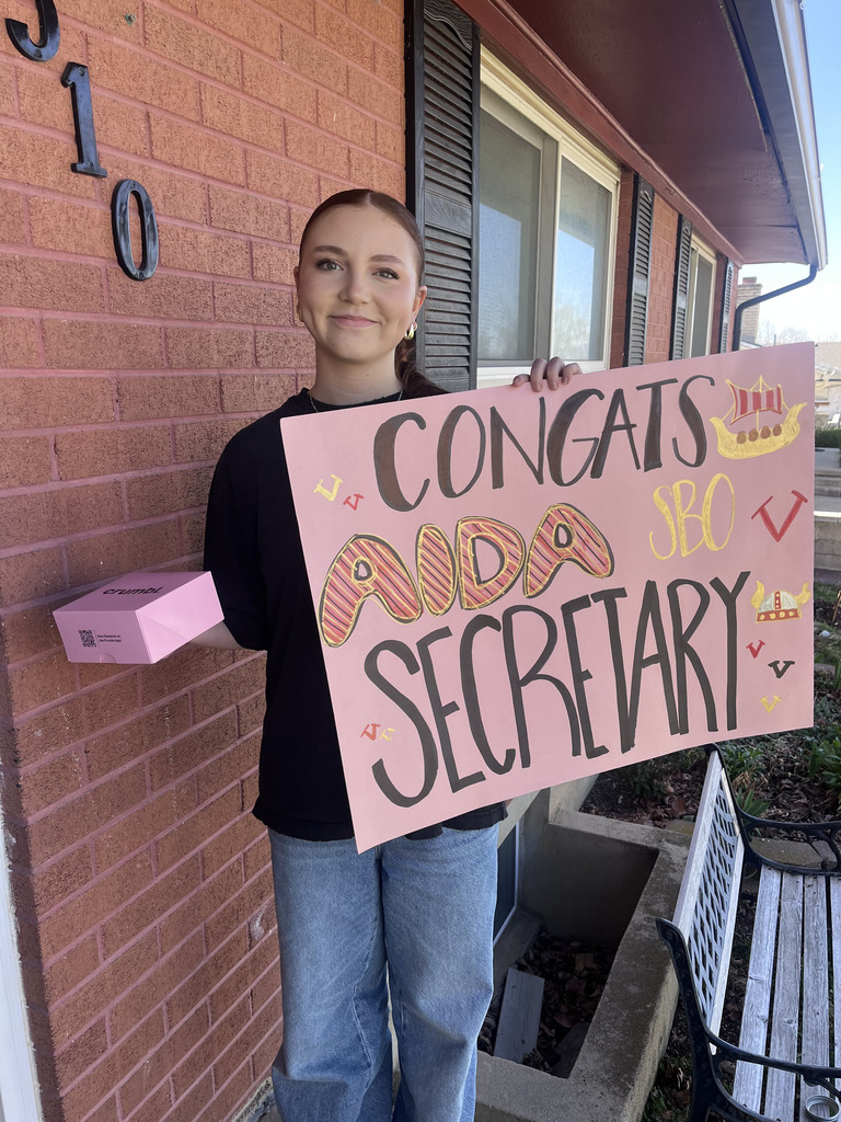 A smiling young person stands on a front porch beside a brick wall, holding a large pink handmade sign that reads “Congrats Aida Secretary,” with “SBO,” Viking drawings, and small decorative letters. They’re wearing a black shirt and jeans and holding a pink Crumbl box, with windows and a bench visible along the porch.