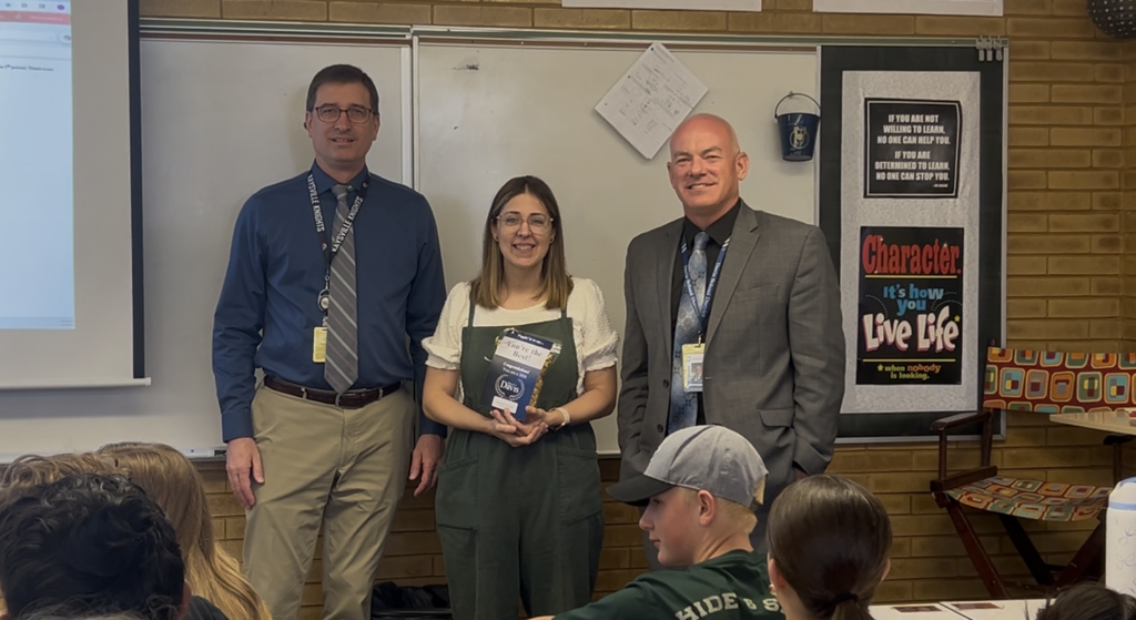 image of teacher principal and director standing in front of a white board. Teacher is holding a best in davis award