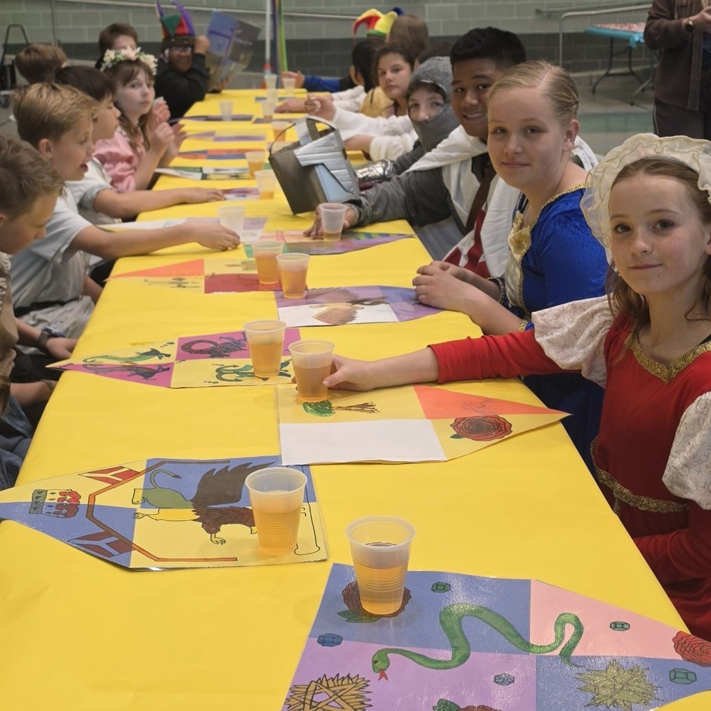 Students in medieval costumes sit along both sides of a long banquet table covered in bright yellow paper, with handmade placemats and plastic cups in front of them during a school feast.
