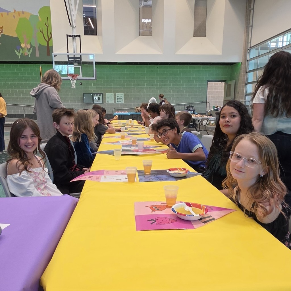 Students sit along both sides of a long yellow-covered banquet table in the school gym, with several smiling toward the camera beside handmade placemats, cups, and bowls during a medieval feast celebration.
