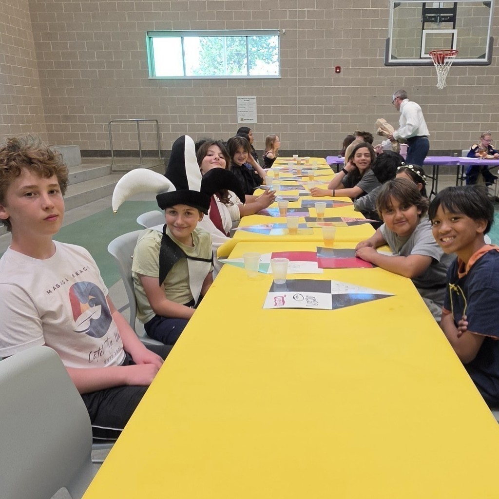 Students sit along a long banquet table covered in bright yellow paper, with cups and handmade placemats in front of them, while several smile toward the camera during a medieval-themed school feast in the gym.