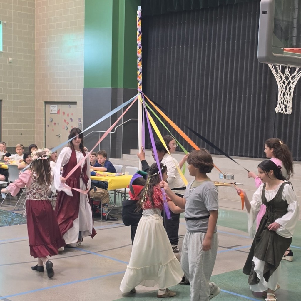 Students in medieval-style costumes hold colorful ribbons and dance around a maypole in the school gym while classmates watch from nearby tables.