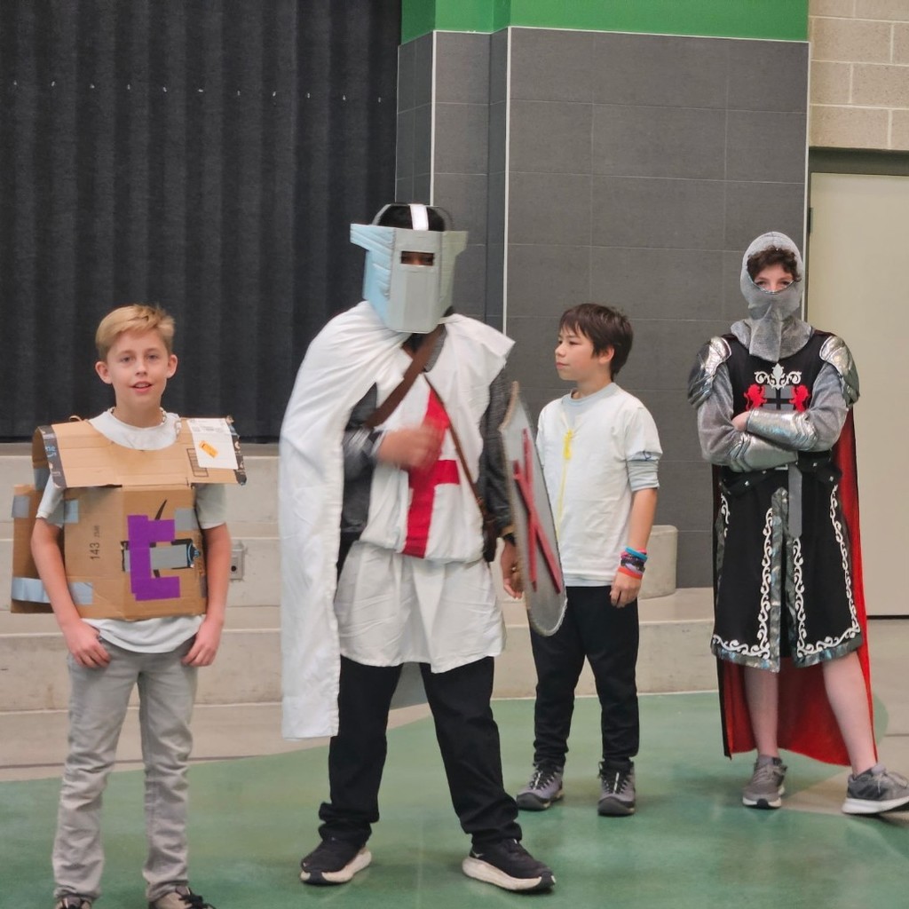 Four students stand together in costume during a medieval-themed school event, including cardboard armor, knight helmets, shields, and capes.
