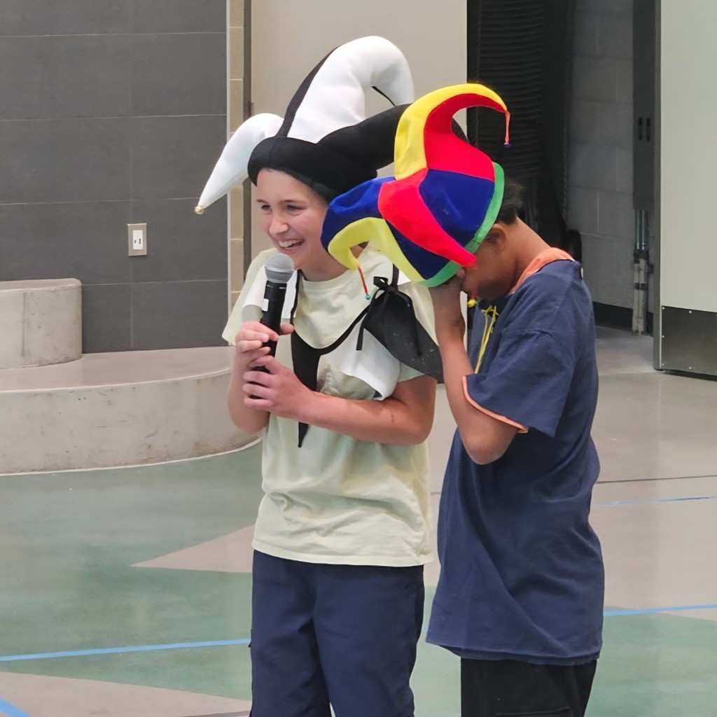 Two students wearing jester hats stand together in the gym, with one smiling and speaking into a microphone while the other covers part of their face.