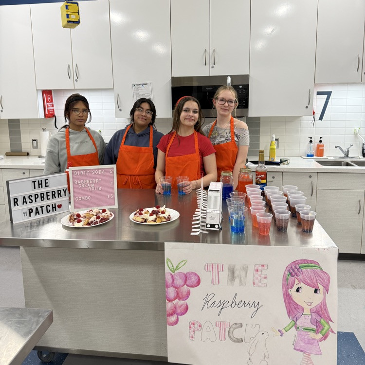 Four students in orange aprons stand behind a table in a classroom kitchen with raspberry-themed desserts, drinks, and a sign that says “The Raspberry Patch.”