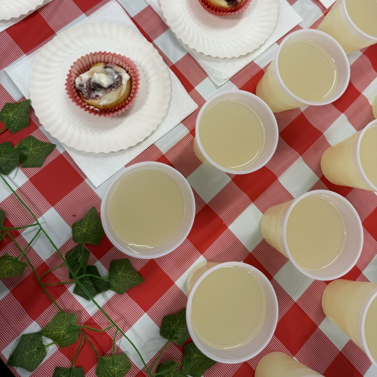 Cinnamon rolls on white plates with frosting and cups of a light-colored drink arranged on a red checkered tablecloth.