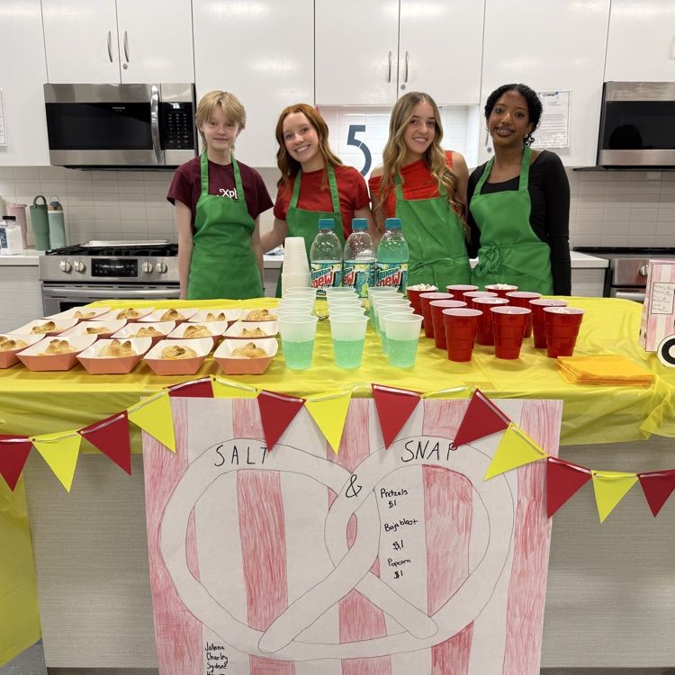 Four students in green aprons stand behind a table in a classroom kitchen selling pretzels, popcorn, and drinks, with red cups, green soda, and a red-and-yellow decorated snack stand display.