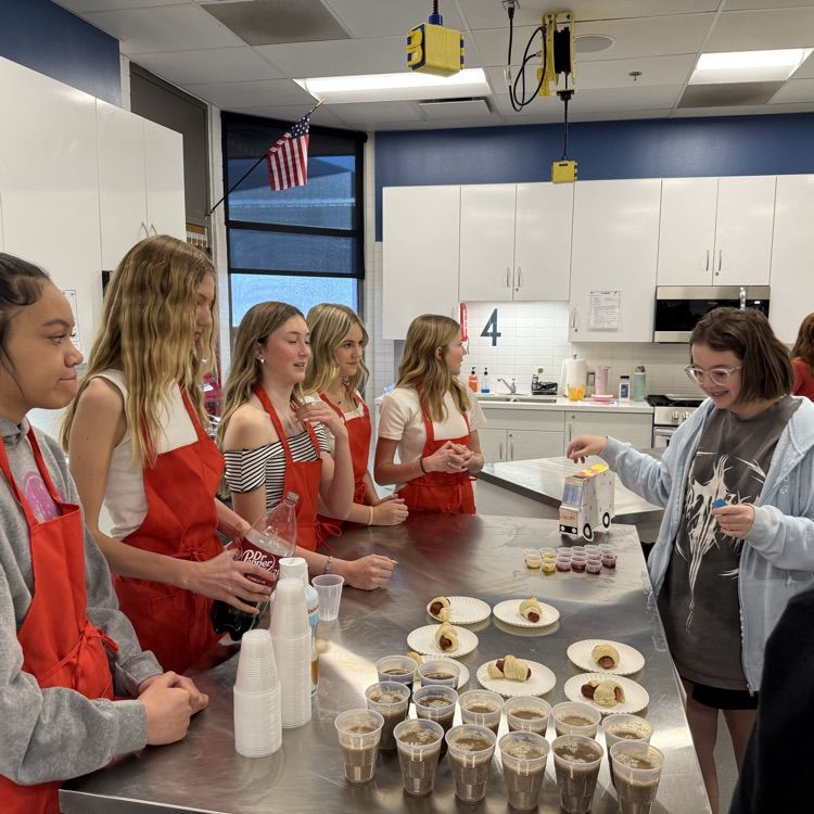 Students in red aprons stand around a table in a classroom kitchen serving hot dog snacks and drinks while another student pays at a small stand, with cups and food arranged on the table.