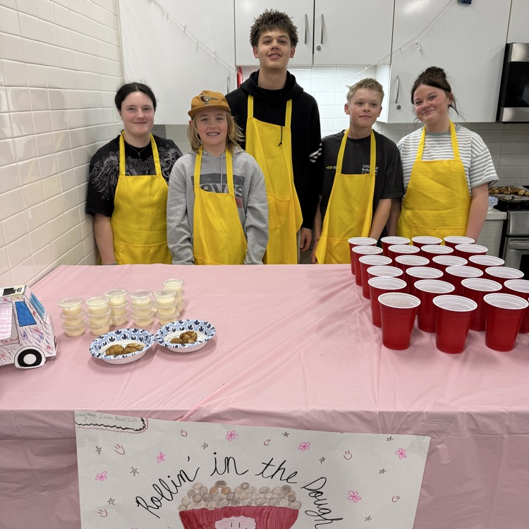 Five students in yellow aprons stand behind a table in a classroom kitchen with cookies, small dessert cups, and rows of red drinks, with a sign that says “Rollin in the Dough.”