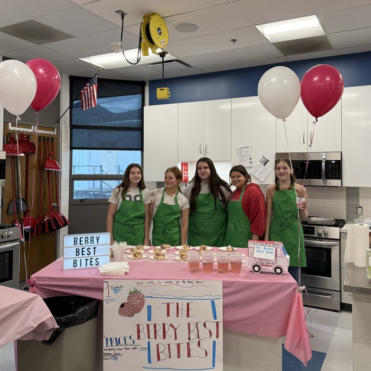Five students in green aprons stand behind a table in a classroom kitchen decorated with pink tablecloths and balloons, serving berry-themed desserts and drinks with a sign that says “The Berry Best Bites.”