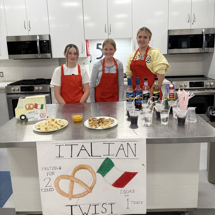 Three students in red aprons stand behind a table in a classroom kitchen with pretzels and Italian soda drinks, with a sign that says “Italian Twist.”