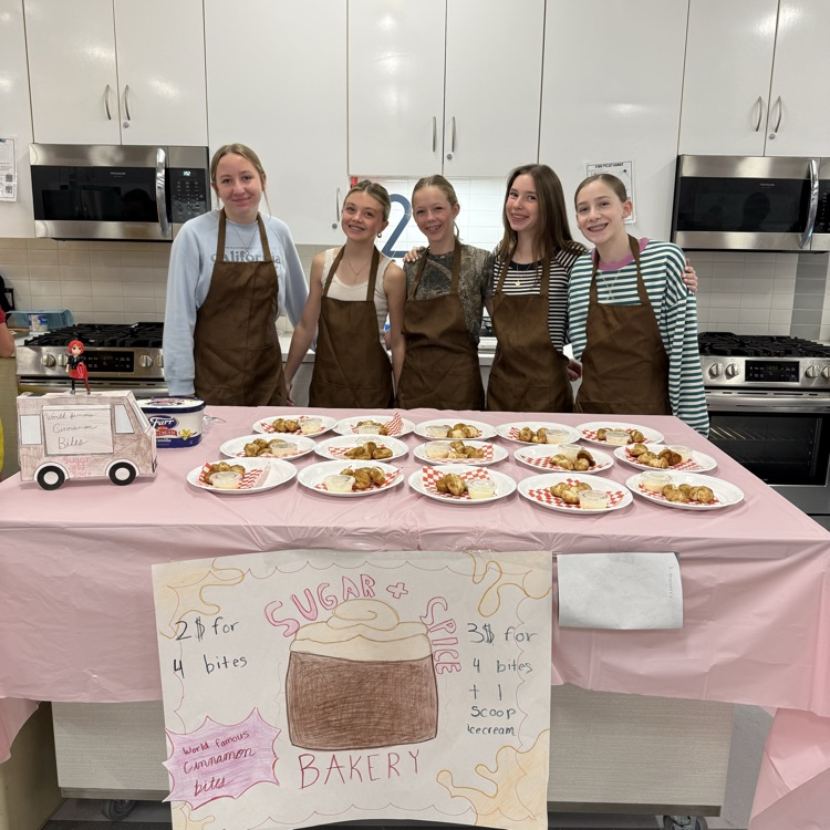 Five students in brown aprons stand behind a table in a classroom kitchen with plates of cinnamon bites and dipping sauce, with a sign that says “Sugar & Spice Bakery.”