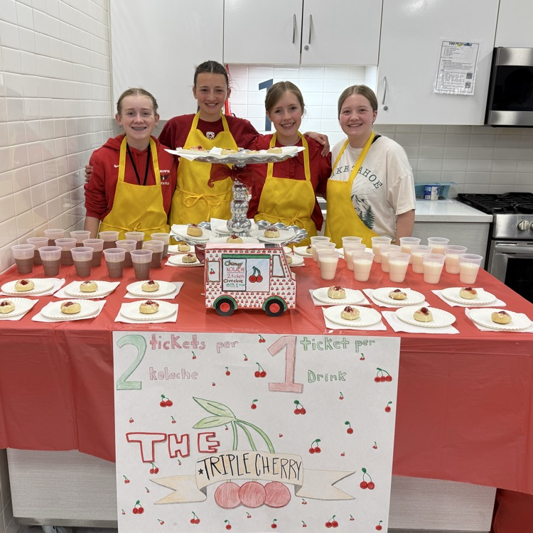 Four students in yellow aprons stand behind a table in a classroom kitchen with kolaches and drinks, with a red tablecloth and a sign that says “The Triple Cherry.”