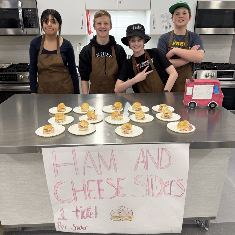 Four students in brown aprons stand behind a table in a classroom kitchen with ham and cheese sliders on plates and a sign that says “Ham and Cheese Sliders – 1 ticket per slider.”