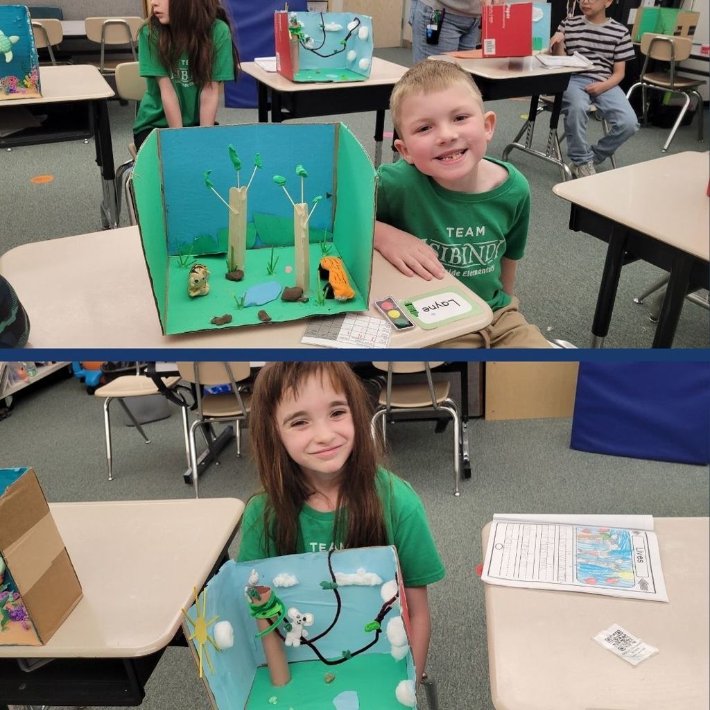 Students sitting next to their animal habitats created using clay, paper, and art supplies.