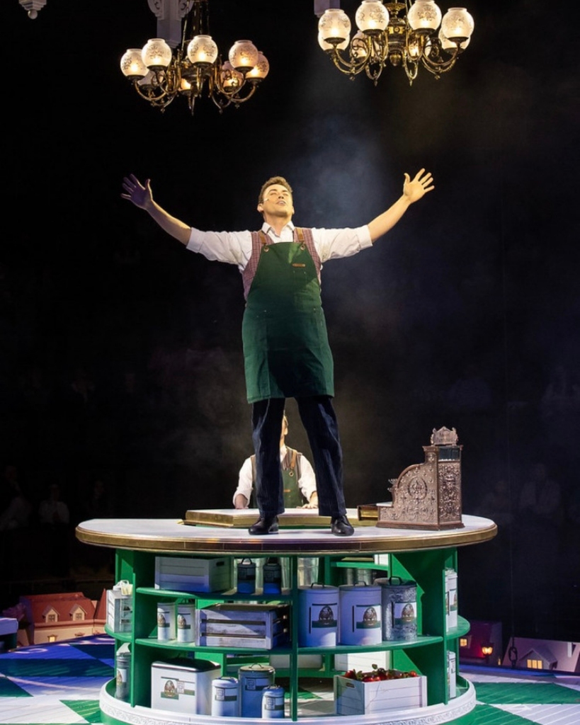 An adult in a green apron standing on a table sings. 
