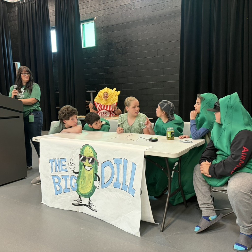 Student team The Big Dill sits at a table during Bountiful Elementary’s student vs. teacher Book Battle assembly, with several students dressed in green pickle costumes while a staff member stands at a podium nearby. A hand-drawn team banner featuring a cartoon pickle and the words “The Big Dill” hangs across the front of the table, and a teacher in a fries costume stands in the background on the school stage.