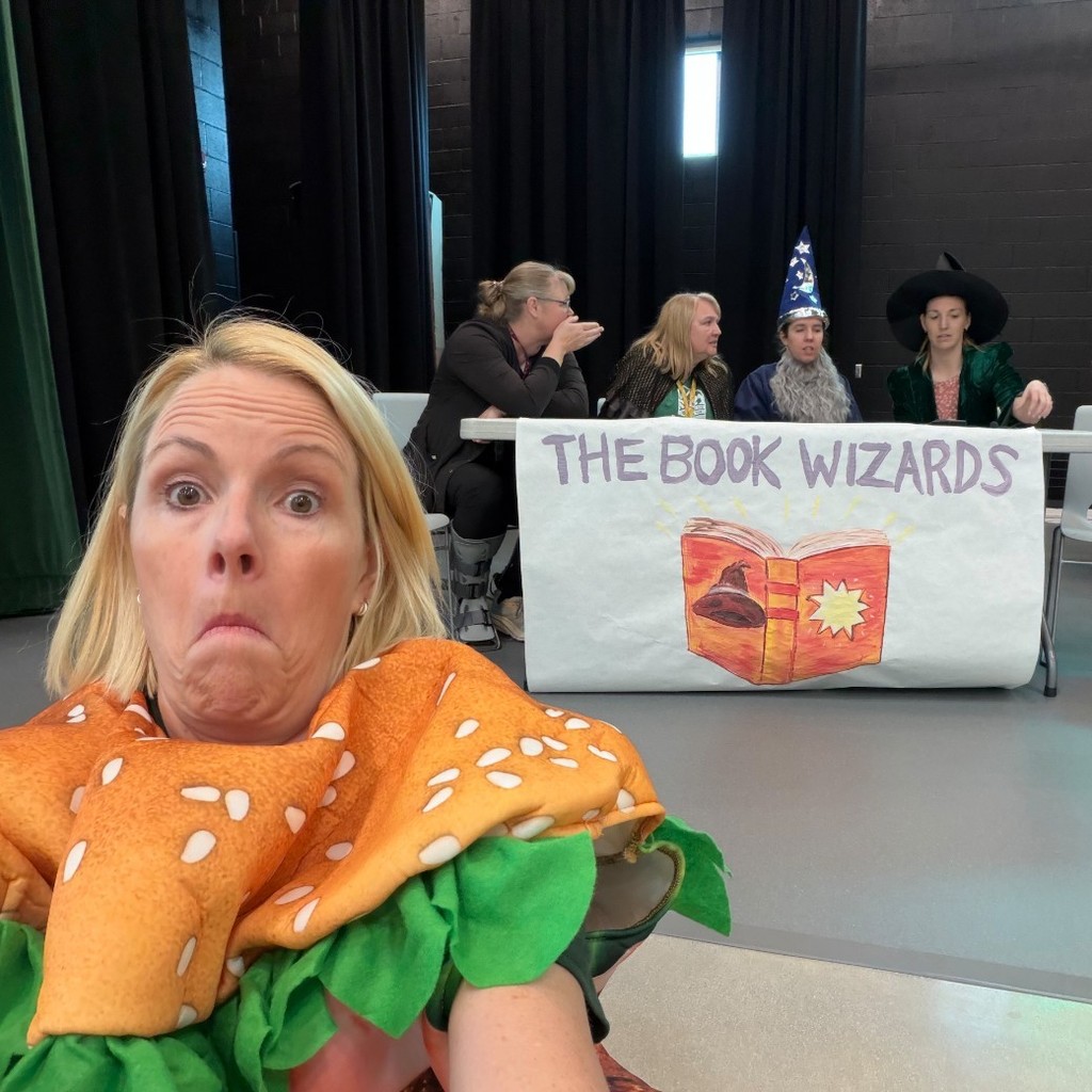 Staff team The Book Wizards sits on stage during Bountiful Elementary’s student vs. teacher Book Battle assembly, with teachers dressed in playful costumes including a wizard hat, witch hat, fries costume, and a hamburger costume in the foreground. A team banner reading “The Book Wizards” with a hand-drawn open book hangs across the front of the table during the school reading competition.