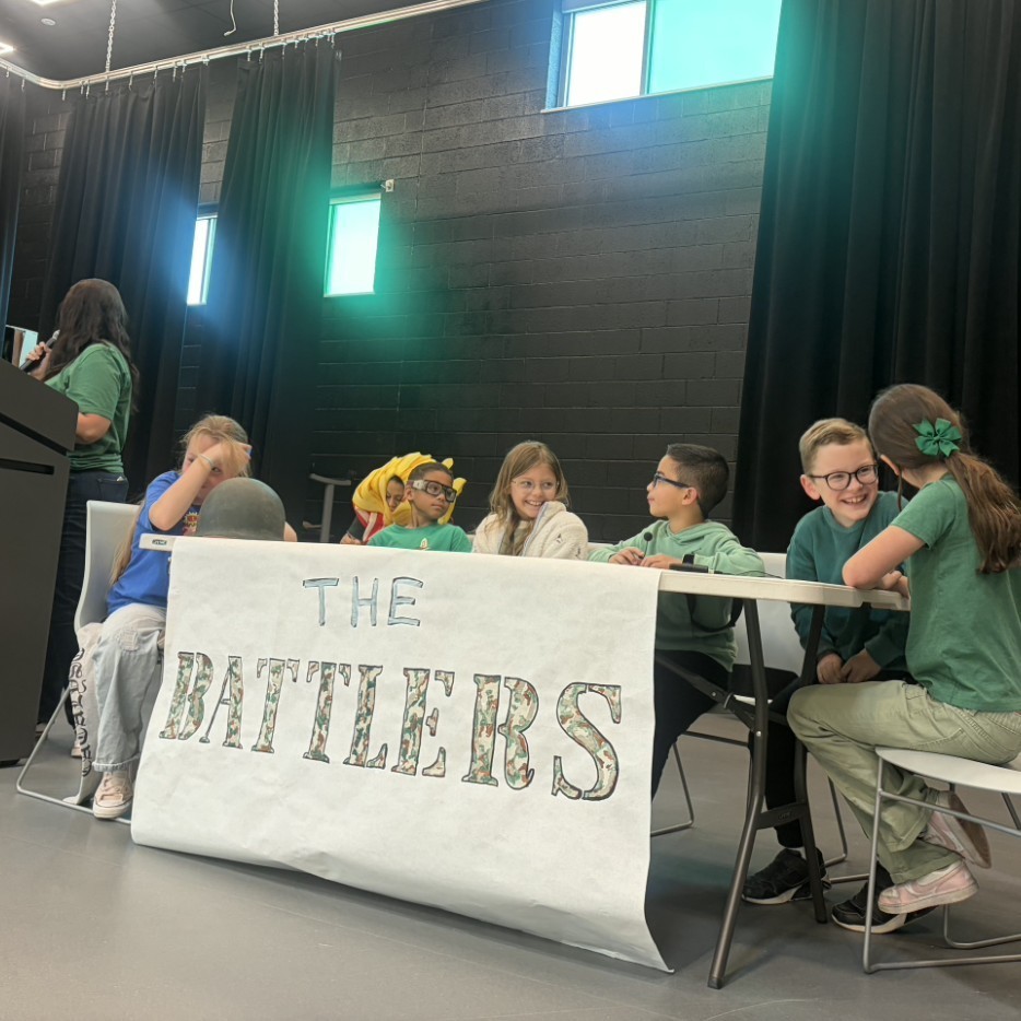 Student team The Battlers sits on stage during Bountiful Elementary’s Book Battle assembly, smiling and talking together behind a hand-lettered team banner. A staff member stands at the podium beside the team as students prepare for the school’s student vs. teacher reading competition.