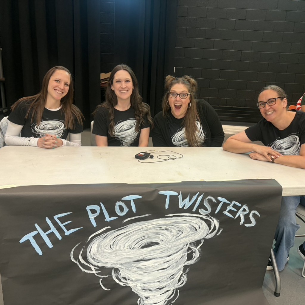 Teacher team The Plot Twisters poses on stage at Bountiful Elementary’s student vs. teacher Book Battle assembly, smiling behind a black team banner painted with a large white tornado. Four staff members wear matching shirts with tornado graphics as they sit at the competition table.