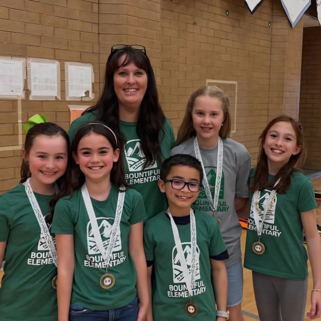Mrs. Berge smiles with five Bountiful Elementary Book Battle students wearing matching green school shirts and medals in the school gym.