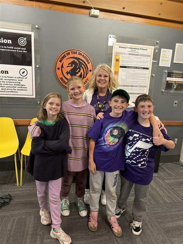 A group of four smiling elementary students and one adult woman standing together in a school hallway in front of a "Home of the Dragons" school seal. They are all wearing various shades of purple clothing, including a purple tie-dye shirt and a purple Utah Jazz jersey, to celebrate Purple Up day for military children.