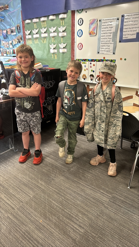 Three elementary-aged students standing in a classroom for Camo Day. On the left, a boy in a mullet and camo shorts crosses his arms confidently. In the center, a boy in a Boba Fett t-shirt and ripped camo pants smiles. On the right, a girl wears an oversized Air Force camouflage uniform jacket and matching cap, smiling at the camera.