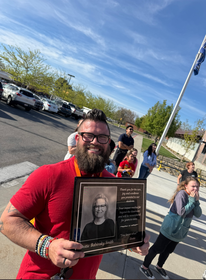 A close-up of a staff member smiling while holding a black memorial plaque for Dottie Babinsky Smith. The plaque includes her photo and a quote that reads, "Thank you for the care, joy and excellence you provided to our students. Your impact reaches far beyond the classroom and will be remembered for years to come. 'You know what?' —Dottie.