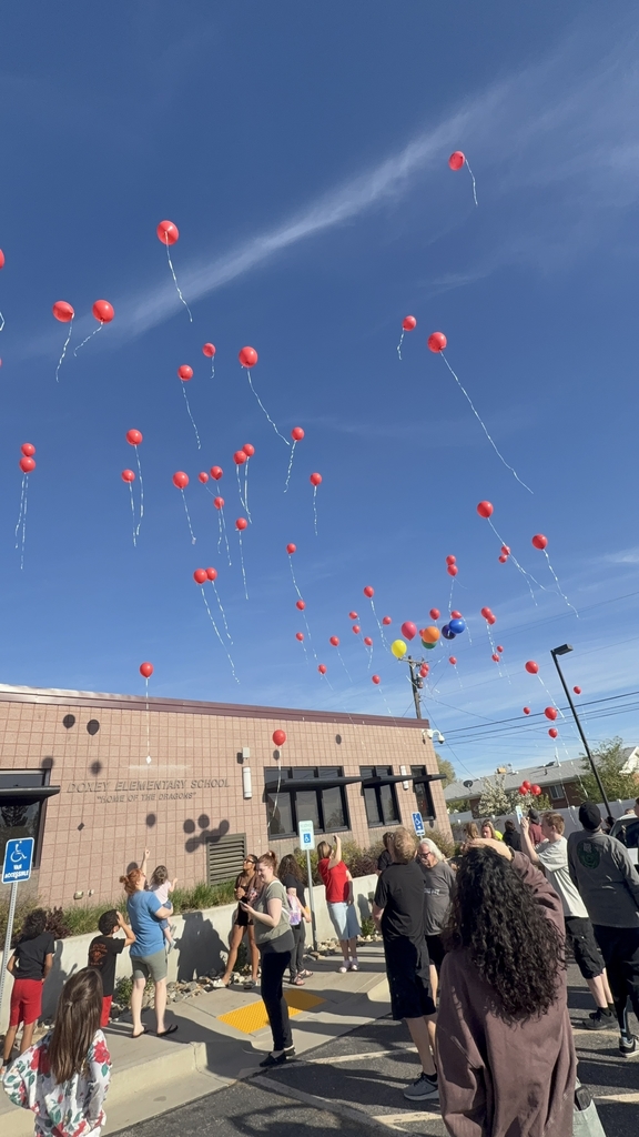 A moving view from the school parking lot showing a diverse crowd of people of all ages looking upward as dozens of her favorite bright red balloons drift into a clear blue sky. The brick exterior of Doxey Elementary School, "Home of the Dragons," is visible in the background under a warm sun.