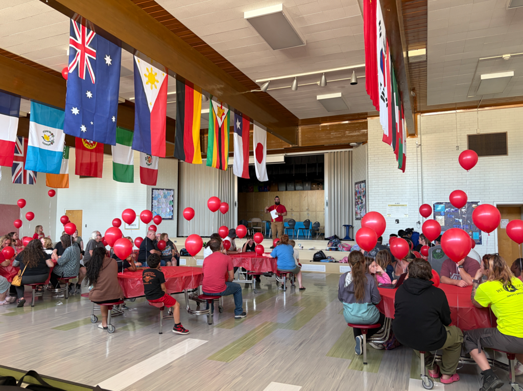 A wide shot of the Doxey Elementary cafeteria filled with students, staff, and families seated at tables with red tablecloths. Each table has several red balloons floating above it. A row of colorful international flags spans the length of the ceiling, and a speaker stands on the stage in the distance.