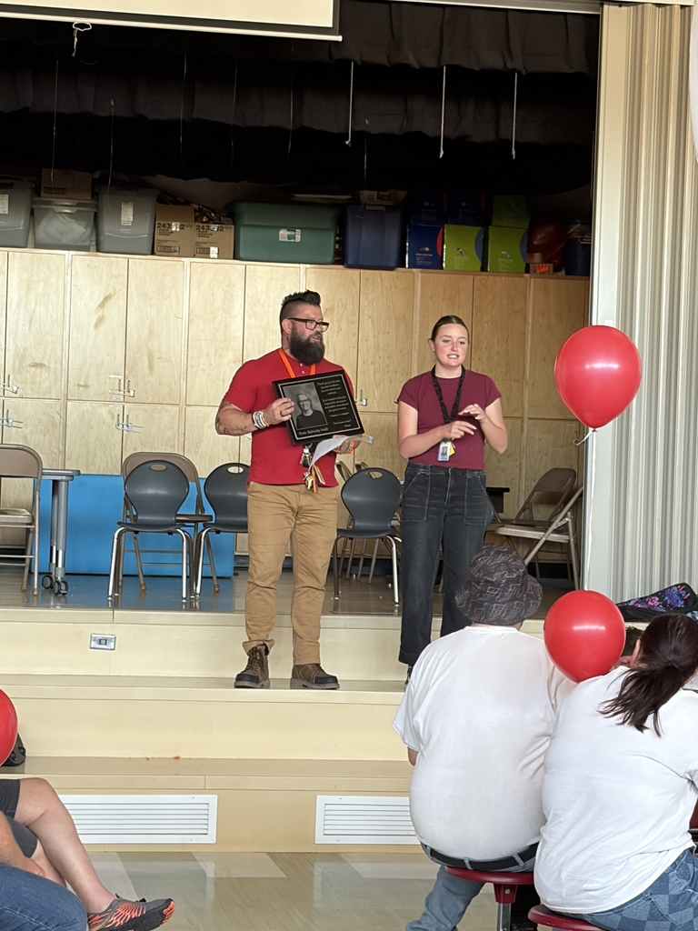 Two staff members stand on the school stage during the memorial service. One man with a beard and glasses holds a memorial plaque featuring a portrait of Dottie, while a woman speaks to the gathered audience. A single red balloon is visible to the right