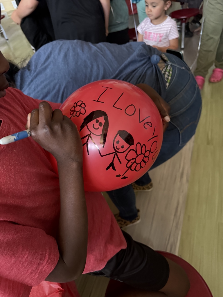 A close-up of a student using a marker to draw a heartfelt message on a red balloon. The drawing shows two stick figures holding hands next to flowers, with the words "I love you" written across the top of the balloon.