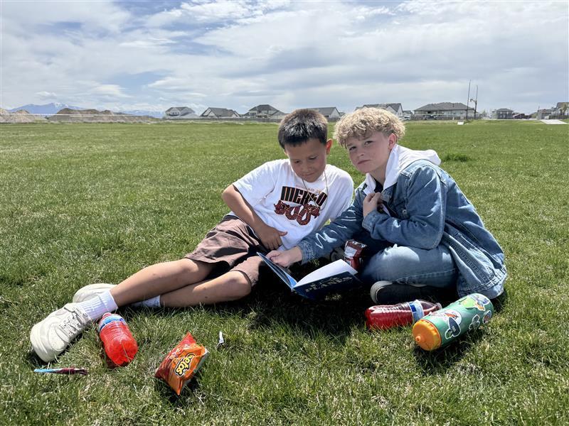two boys reading a book outside on the grass