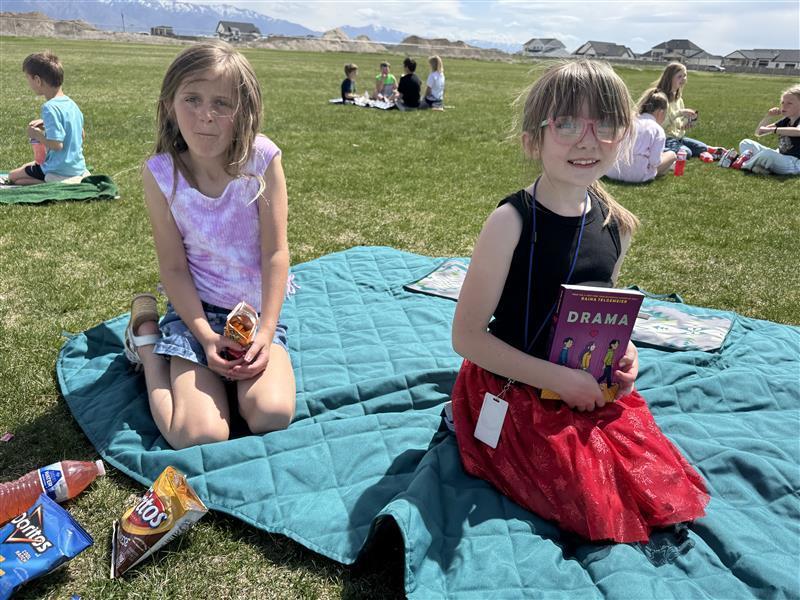 two girls sitting on a blue blanket in grass with books and chips