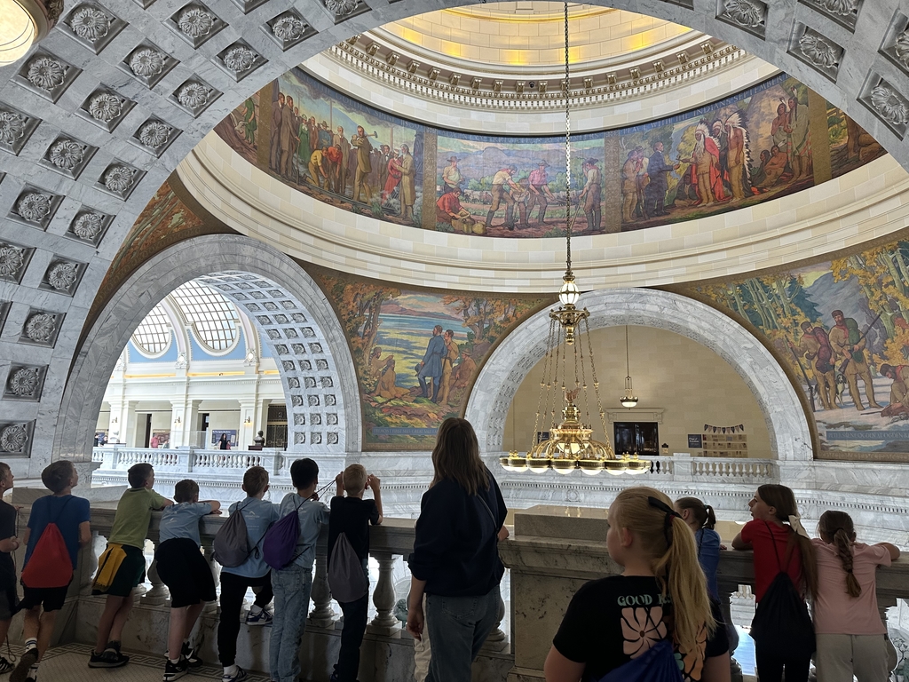 group of kids and a women looking over a balcony with large gold chandelier and color paintings on the walls