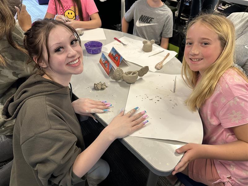 a girl in a hoodie and a young girl in pink smile at the camera with a clay pot with fins in the background