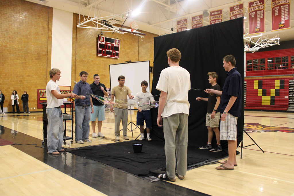A group of students stand in a circle inside a school gym, each holding a rope to create a large soap bubble stretched between them. A black backdrop and projector screen are set up behind them, and a small bucket sits on the floor in the center of the circle. Banners and a scoreboard hang on the gym walls in the background.