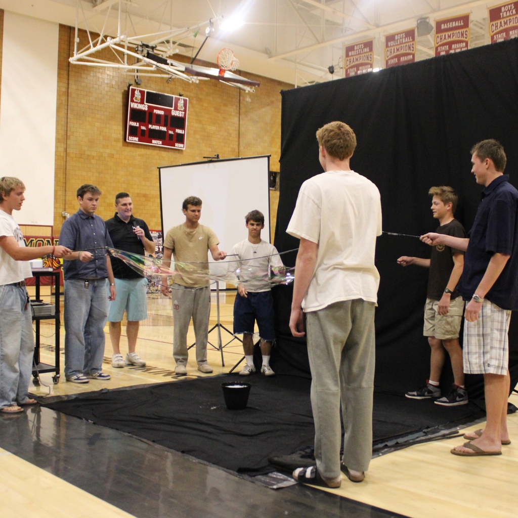 A group of students stands in a school gymnasium holding rods to create a large soap bubble during a bubble assembly demonstration, with a black backdrop and other students watching in the background.