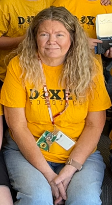 A woman with long blonde hair wearing a bright yellow "Doxey Robotics" t-shirt and a school lanyard. She is sitting with her hands clasped, smiling gently during a team event.