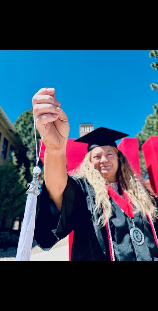 A close-up, low-angle shot of a graduate in a black cap and gown holding up a white tassel with a silver "26" charm toward the camera. In the blurred background, she has a proud smile, standing in front of a bright red "SUU" sign under a clear blue sky.