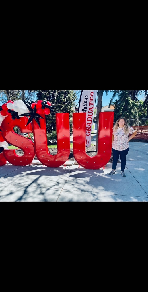 A graduate standing next to large, red marquee-style letters that spell "SUU," decorated with black, white, and red balloons. A banner in the background reads "Congratulations Graduates," and the scene is set outdoors on a sunny campus day.