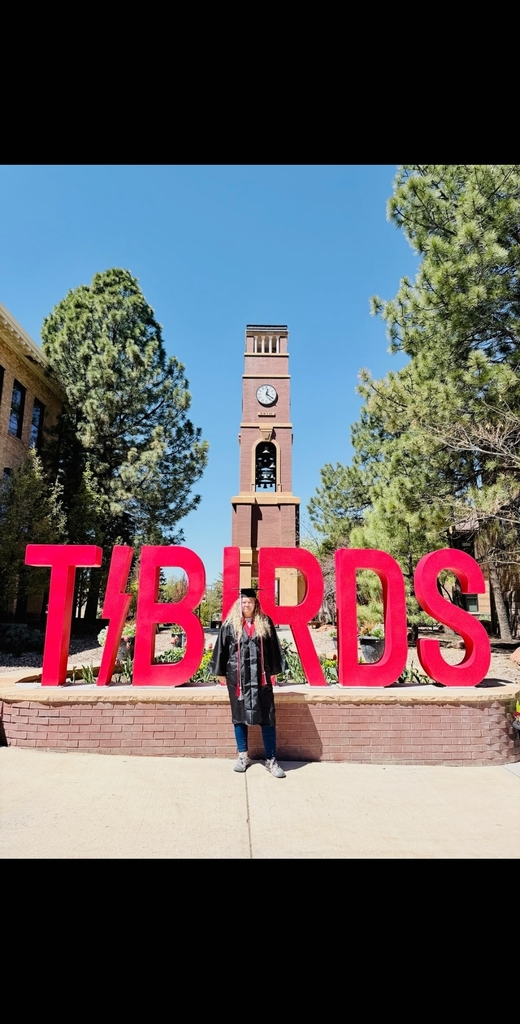 A wide shot of a graduate in a black cap and gown standing behind large, bright red letters that spell "T-BIRDS." In the background, the Southern Utah University brick bell tower rises into a bright blue sky, framed by lush green pine trees.