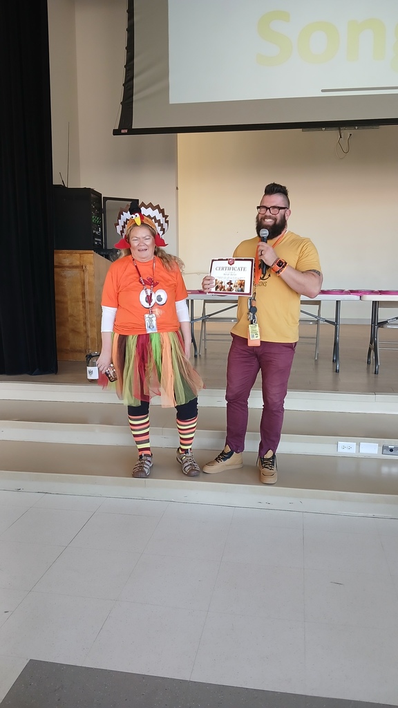 A woman dressed in a festive turkey costume, including a themed hat and a colorful tutu, standing on a school stage. Next to her, a man in a yellow shirt holds a microphone and a certificate, presenting it to her during a school assembly.