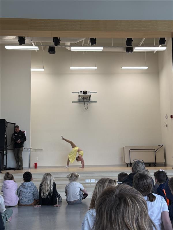  A student performer in a yellow dance costume performs a backbend with one leg extended upward on a wooden school stage. Several students sit on the floor in the foreground, watching the performance from behind. An adult stands to the side of the stage near some equipment, and a projector is mounted on the plain back wall under the stage lights.