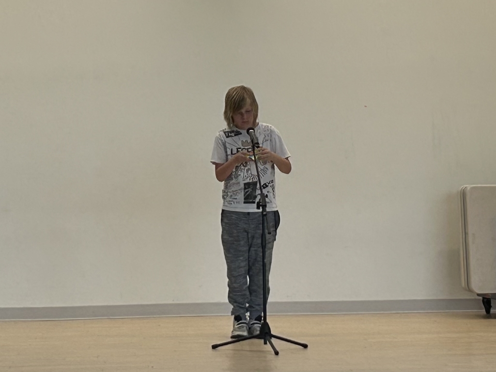 A student performer stands center stage at a microphone, looking down as they solve a triangular Rubik's pyramid (Pyraminx) with their hands. They are wearing a white graphic t-shirt with a "Legend" design and grey patterned pants. The background is a large, plain cream-colored wall, and the stage is made of light-colored wood.